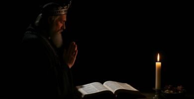 Obraz przedstawiający księdza modlącego się z zamkniętymi oczami obok otwartej Biblii, zapalonej świecy i owoców. W tle znajduje się ciemne, niczym smoła pomieszczenie, w którym nie widać nic poza modlącym się księdzem.