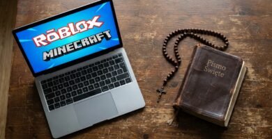 Obraz laptopa na stole z symbolami Roblox i Minecraft na ekranie; obok leży stara Biblia i różaniec.